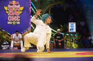 Dancer with blue hair performs in a breakdancing competition, wearing a white tracksuit, at the Red Bull BC One World Final event