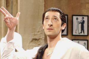 Adrien Brody in a white shirt and black pants strikes a dramatic pose in a room with framed photos and wooden chairs