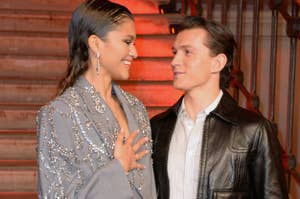 Zendaya and Tom Holland smile at each other. One wears a jeweled blazer, the other a leather jacket. The setting appears elegant and formal